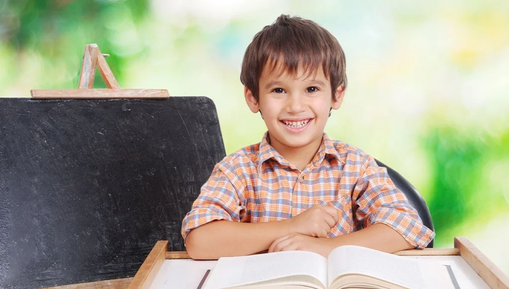 Young boy smiling while sitting beside a chalkboard and a book infront of him