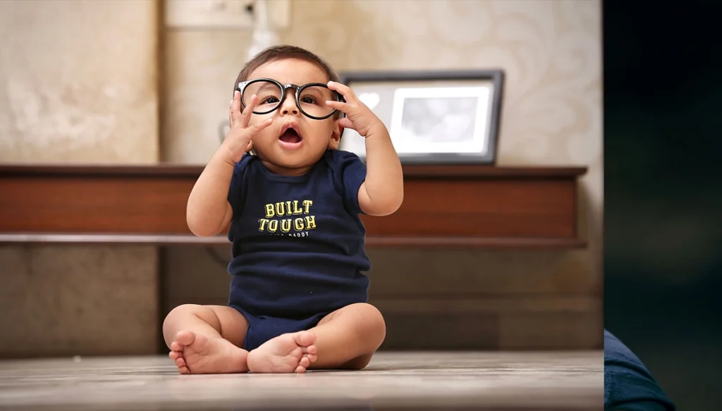 baby wearing oversized glasses, sitting indoors with playful expression.