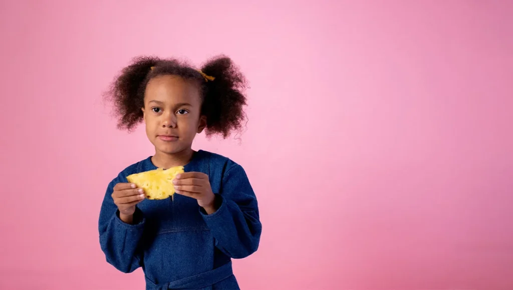 Child in blue dress holding pineapple slice with playful expressions on a pink background.